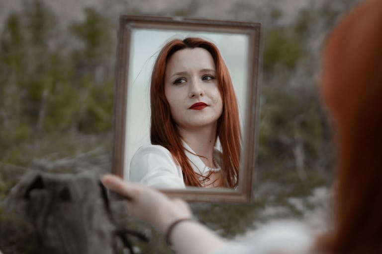 A young redhead woman holding a mirror outdoors, showcasing her reflection with red lips.